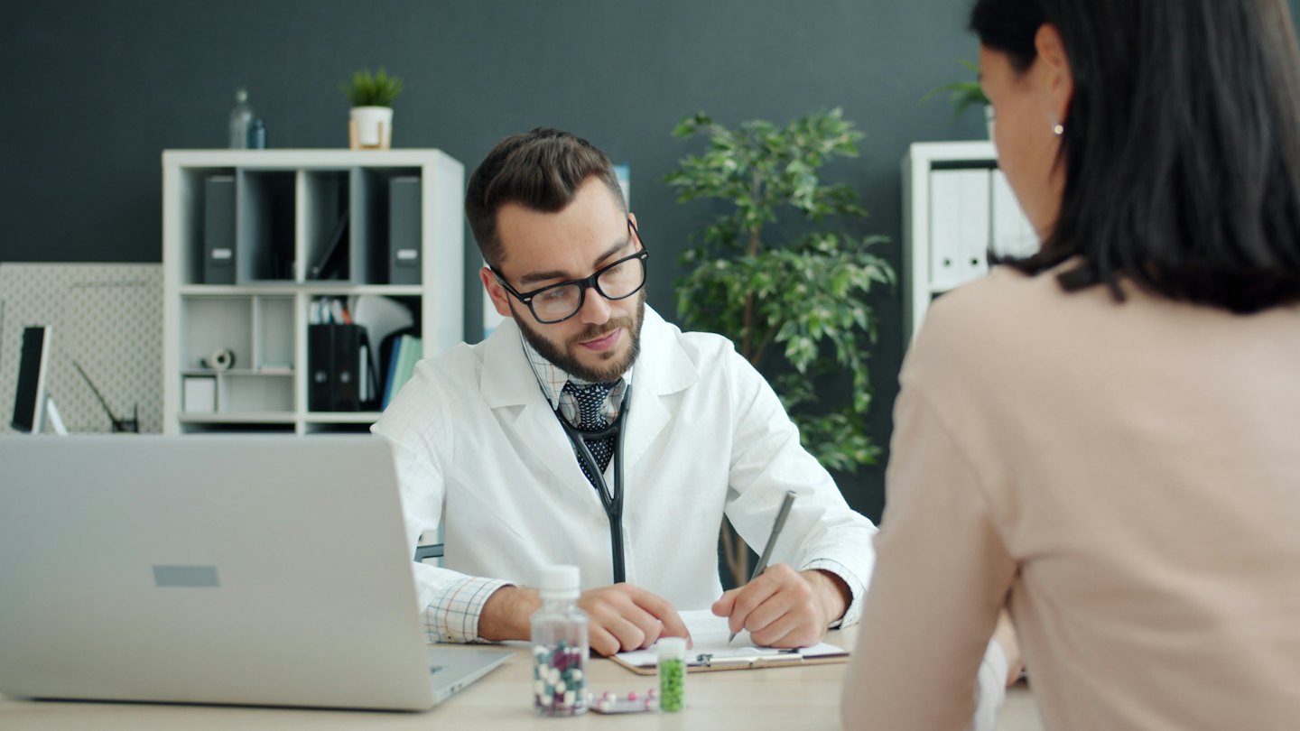 Läkare skriver på clipboard under patientkonsultation vid skrivbord.